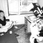 Bärbel Bohley im Schneidersitz auf einem Polstermöbel, neben sich einen aschenbecher und Zigaretten. Im Vordergrund ein Teil einer Kamera, die sie filmt.