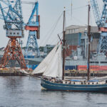 Ein Segelboot zieht an den Kieler Industrieanlagen vorbei. Im Hintergrund: ein dort liegendes Marineschiff.