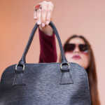 Eine junge Frau mit Sonnenbrille und langen dunklen Haaren hält eine schwarze Handtasche in Richtung auf die Kamera hoch.